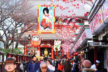 Eine belebte Straße in Japan zu Hatsumōde, die mit Laternen, roten und weißen Ornamenten und großen Schildern mit traditionellen Kunstwerken, einer Daruma-Puppe, einer Glückskatze und Geschäften auf beiden Seiten geschmückt ist.