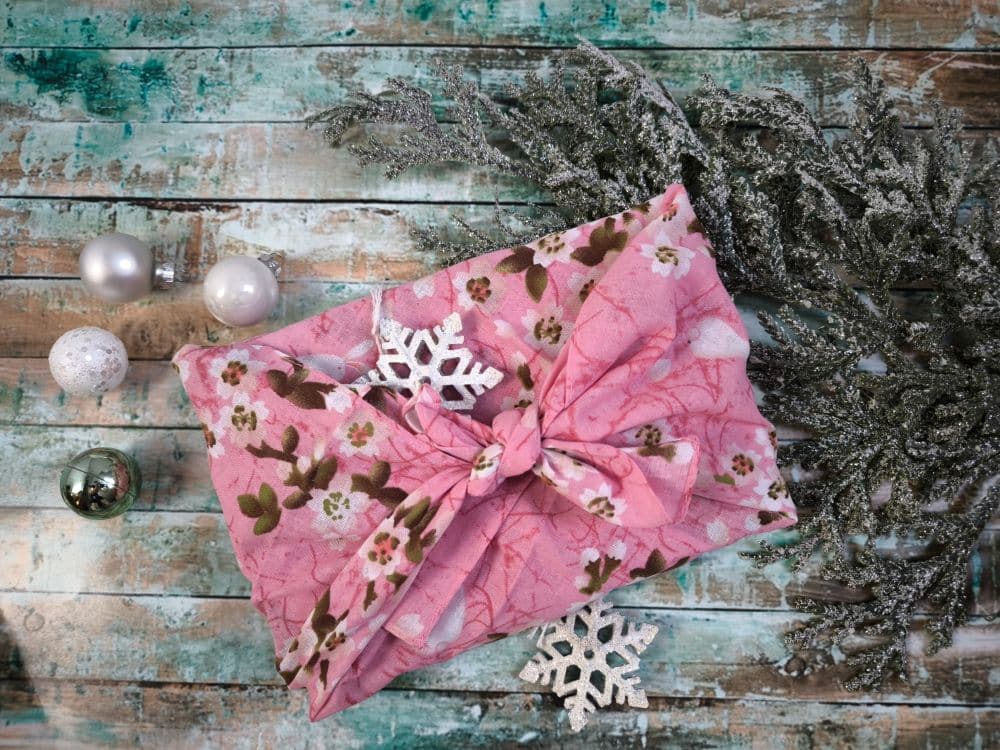 Ein in rosa Blumentuch mit Knoten verpacktes Furoshiki-Geschenk liegt auf einer rustikalen Holzfläche, die mit weißen Schneeflocken, silbernen und weißen Ornamenten und gefrostetem Grün verziert ist.