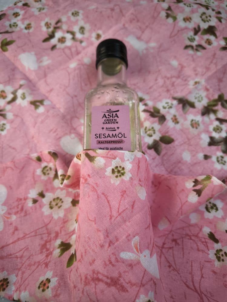 Eine Flasche Sesamöl von ASIA Garden ist teilweise in ein rosafarbenes, blumengemustertes Furoshiki-Tuch mit weißen Häschen und grünen Blättern eingewickelt. Das Etikett der Flasche ist vor demselben rosa Stoffhintergrund zu sehen.