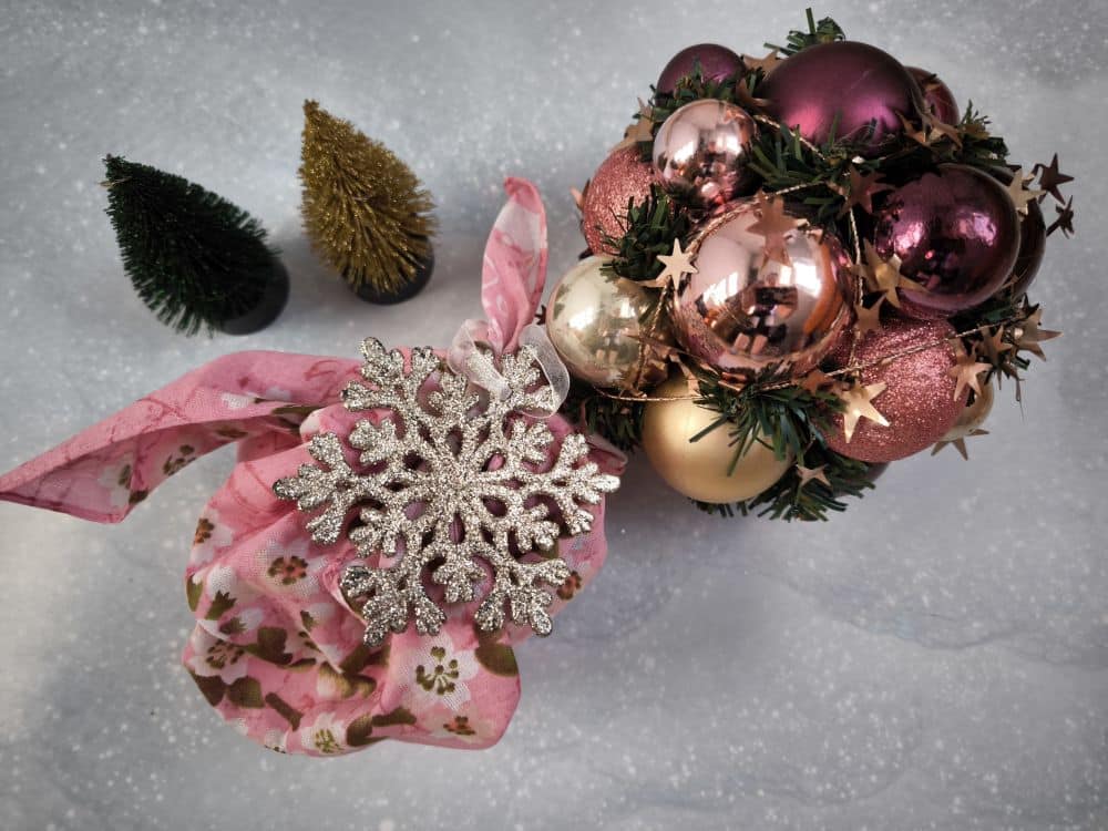 Ein festliches Arrangement mit rosafarbenen und goldenen Ornamenten, einer dekorativen Schneeflocke und einer goldenen Sternengirlande auf Grünzeug, alles eingewickelt in ein rosafarbenes Furoshiki-Blumentuch, steht neben zwei kleinen künstlichen Bäumen auf einer grauen Fläche.