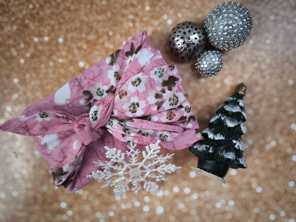 Ein mit rosa Blumen verpacktes Furoshiki-Geschenk steht neben einem kleinen, schneebedeckten Weihnachtsbaum, funkelndem Silberschmuck und einer weißen Schneeflockendekoration auf einem glitzernden Goldhintergrund.