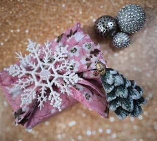 Ein in rosa Blumenstoff verpacktes Furoshiki-Geschenk, das mit einer weißen Schneeflocke und einem kleinen Tannenzapfen-Weihnachtsbaum verziert ist, steht auf einem glitzernden goldenen Hintergrund neben zwei silbernen, mit Perlen verzierten Kugeln.