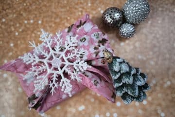 Ein in rosa Blumenstoff verpacktes Furoshiki-Geschenk, das mit einer weißen Schneeflocke und einem kleinen Tannenzapfen-Weihnachtsbaum verziert ist, steht auf einem glitzernden goldenen Hintergrund neben zwei silbernen, mit Perlen verzierten Kugeln.
