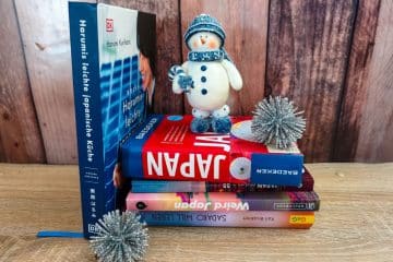 Ein Stapel Bücher über Japan wird von einer kleinen Schneemann-Figur mit Mütze und Schal gekrönt. Zwei silberne Bommeln ruhen neben den Büchern auf einer hölzernen Unterlage und bilden charmante Geschenkideen für Japan-Fans.