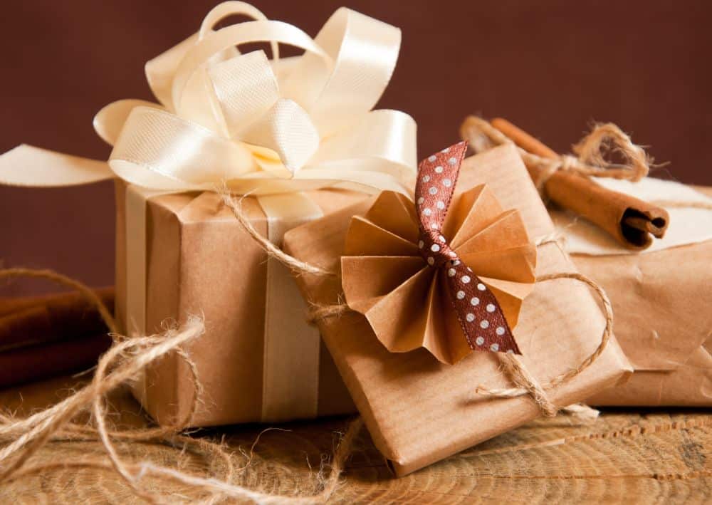 Zwei in braunes Papier eingewickelte Geschenke, perfekt für O-seibo, sind mit Zwillingen verziert - eines mit einem cremefarbenen Band, das andere mit einer Papierrosette und einer gepunkteten Schleife. Zimtstangen liegen in der Nähe auf einer Holzfläche.