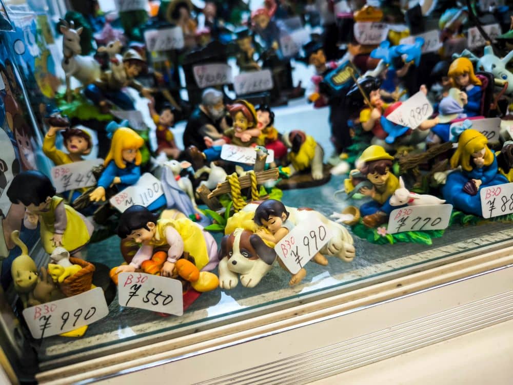 Ein Schaufenster, das an den Nakano Broadway erinnert, zeigt bunte Comic- und Anime-Figuren, die jeweils mit Preisen in thailändischen Baht versehen sind. Die Figuren sind dicht aneinandergereiht, so dass ein lebhaftes und dichtes Bild entsteht.