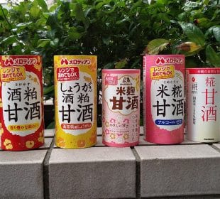 Fünf farbenfrohe Dosen Amazake, das traditionelle süße Reisgetränk, sind mit verschiedenen Etiketten und Mustern auf einer Steinfläche aufgereiht, während das Grün im Hintergrund sanft verschwimmt.