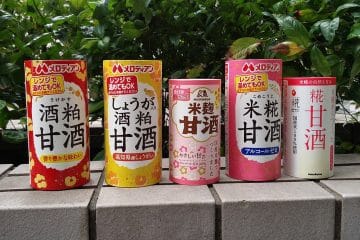 Fünf farbenfrohe Dosen Amazake, das traditionelle süße Reisgetränk, sind mit verschiedenen Etiketten und Mustern auf einer Steinfläche aufgereiht, während das Grün im Hintergrund sanft verschwimmt.