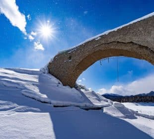 Die Sonne scheint hell am blauen Himmel über dem schneebedeckten Skigebiet und beleuchtet einen alten Steinbogen, der teilweise unter Schnee begraben ist, sowie die fernen Berge am Horizont.