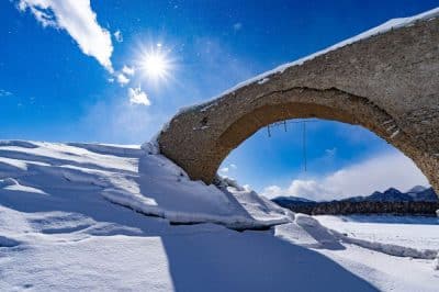 Die Sonne scheint hell am blauen Himmel über dem schneebedeckten Skigebiet und beleuchtet einen alten Steinbogen, der teilweise unter Schnee begraben ist, sowie die fernen Berge am Horizont.