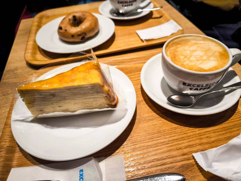 Ein Stück Schichtkuchen, eine Tasse Cappuccino und ein Löffel auf einem weißen Teller liegen auf einem Holztisch im Nakano Broadway. Im Hintergrund stehen auf einem Tablett eine Kaffeetasse, ein Löffel, eine Serviette und ein Schokoladenbrötchen.