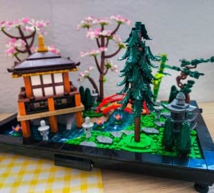 Ein detailliertes LEGO Modell eines japanischen Gartens mit einem kleinen Pavillon, einer hohen Kiefer, einer Steinlaterne, einem Teich mit Seerosen, einer geschwungenen roten Brücke, üppigem Grün und blühenden Kirschbäumen im Hintergrund.