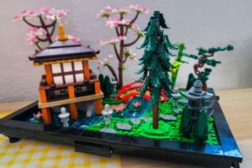 Ein detailliertes LEGO Modell eines japanischen Gartens mit einem kleinen Pavillon, einer hohen Kiefer, einer Steinlaterne, einem Teich mit Seerosen, einer geschwungenen roten Brücke, üppigem Grün und blühenden Kirschbäumen im Hintergrund.