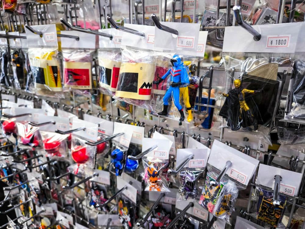 Am Nakano Broadway sind Reihen von bunten Action-Figuren, Spielzeug und Sammlerstücken in durchsichtigen Plastiktüten an Haken ausgestellt. Jede Tüte ist mit einem Preisschild in Yen versehen, das die lebhafte Atmosphäre dieses kultigen japanischen Einkaufsortes widerspiegelt.