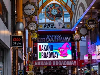 Überdachte Einkaufsstraße mit hell erleuchteten Nakano Broadway-Schildern, einer bunten Zeichentrickfigur auf einem digitalen Bildschirm und einer dekorativen Uhr an der Decke darüber. Menschenmassen flanieren unter der lebhaften Nakano Broadway-Beschilderung.