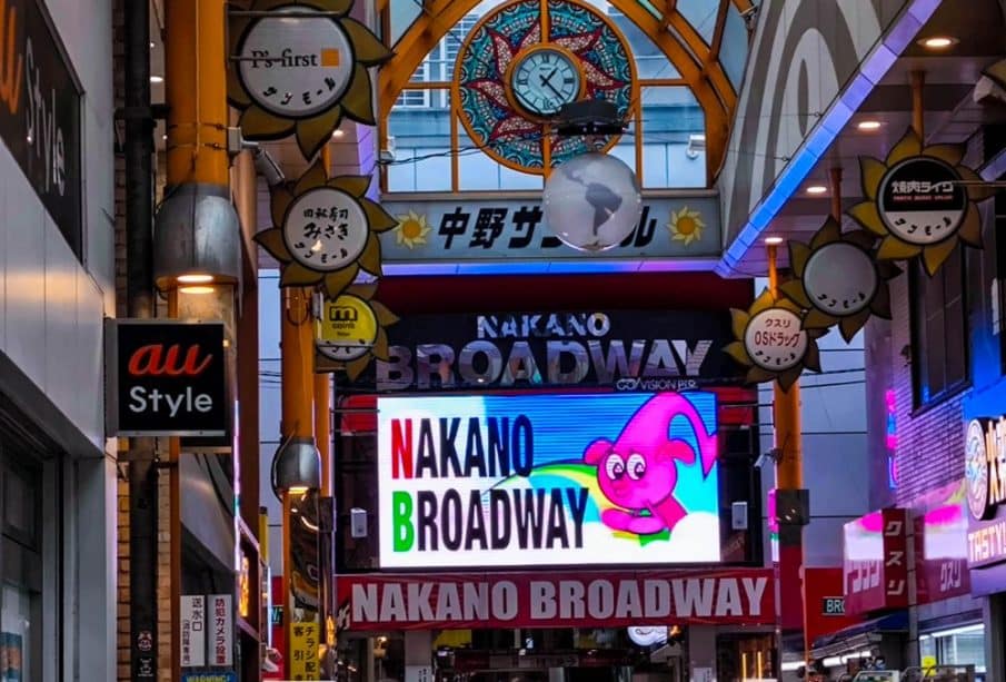 Überdachte Einkaufsstraße mit hell erleuchteten Nakano Broadway-Schildern, einer bunten Zeichentrickfigur auf einem digitalen Bildschirm und einer dekorativen Uhr an der Decke darüber. Menschenmassen flanieren unter der lebhaften Nakano Broadway-Beschilderung.