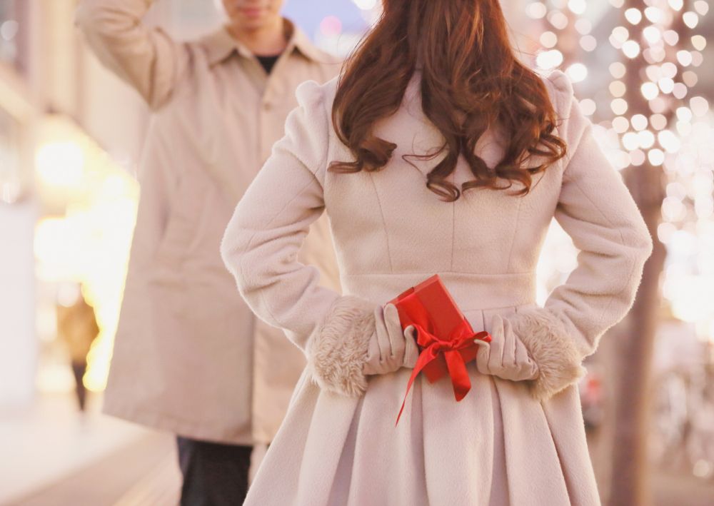 Eine Frau in einem hellen Mantel hält eine kleine rote O-seibo-Geschenkbox hinter ihrem Rücken, während sie einem Mann in einem passenden Mantel auf einer festlichen, beleuchteten Straße gegenübersteht.