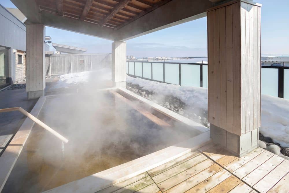 Ein japanisches Onsen-Bad im Freien dünstet unter einem Holzdach, umgeben von Schnee in einem ruhigen Skigebiet, mit einem Milchglaszaun und einer malerischen Aussicht im Hintergrund.