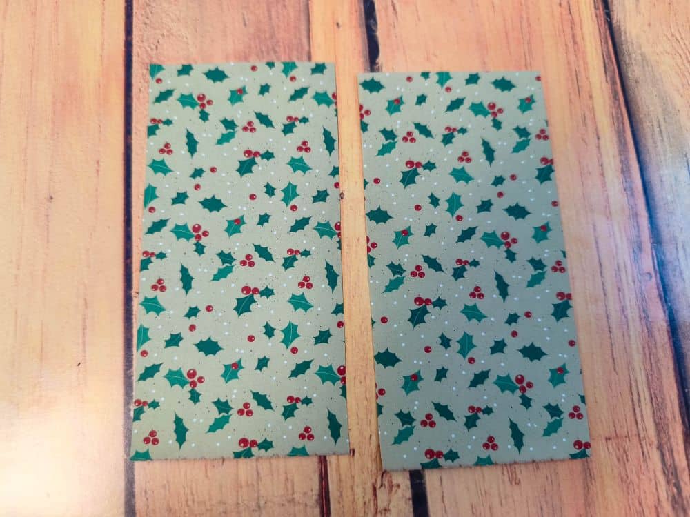 Zwei rechteckige Origamipapierstücke mit einem festlichen Muster aus grünen Stechpalmenblättern und roten Beeren auf hellem Grund, die nebeneinander auf eine Holzfläche gelegt werden.