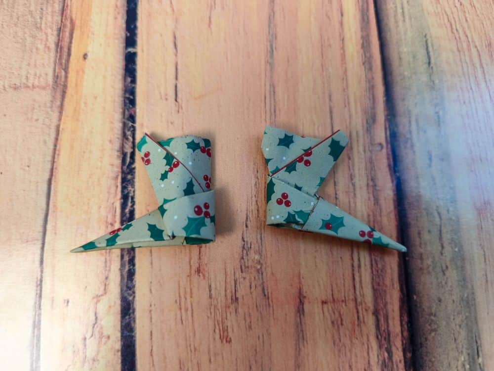 Zwei kleine Stücke gemusterten Origamipapiers, verziert mit Stechpalmenblättern und roten Beeren, werden zu eckigen Formen gefaltet, die an Elfenschuhe erinnern, und auf eine hölzerne Unterlage gelegt.