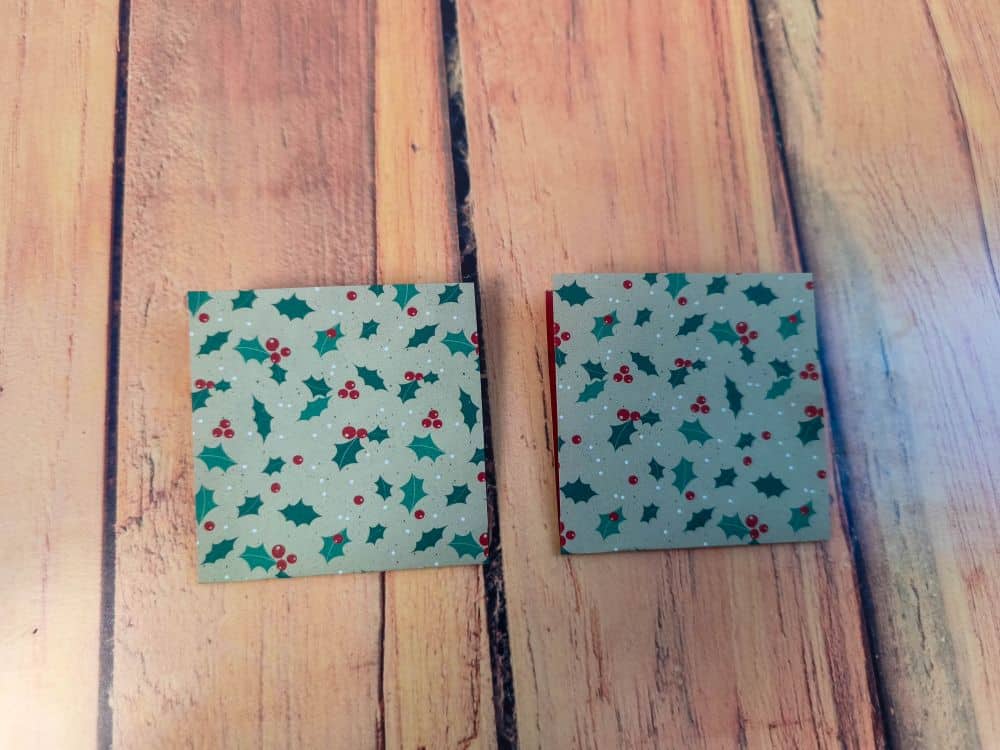 Zwei kleine quadratische Origamipapierstücke mit einem Stechpalmen- und Beerenmuster liegen nebeneinander auf einer Holzfläche.
