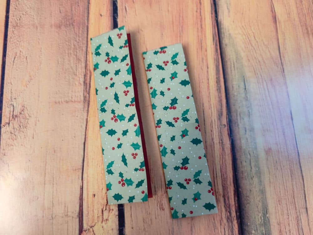 Zwei rechteckige Origamipapierstücke mit einem Muster aus grünen Stechpalmen und roten Beeren werden auf eine Holzfläche gelegt.