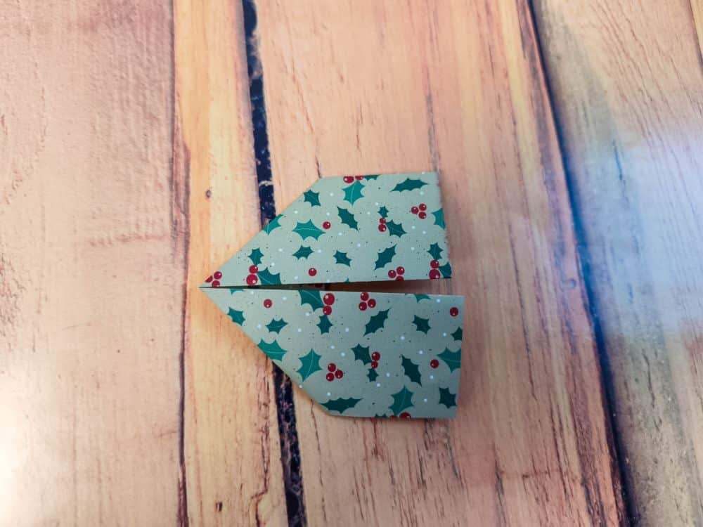 Ein Stück Origamipapier mit einem Muster aus grünen Stechpalmen und roten Beeren wird teilweise auf einer Holzfläche gefaltet, so dass ein Pfeil oder eine Drachenform entsteht, die nach links zeigt.