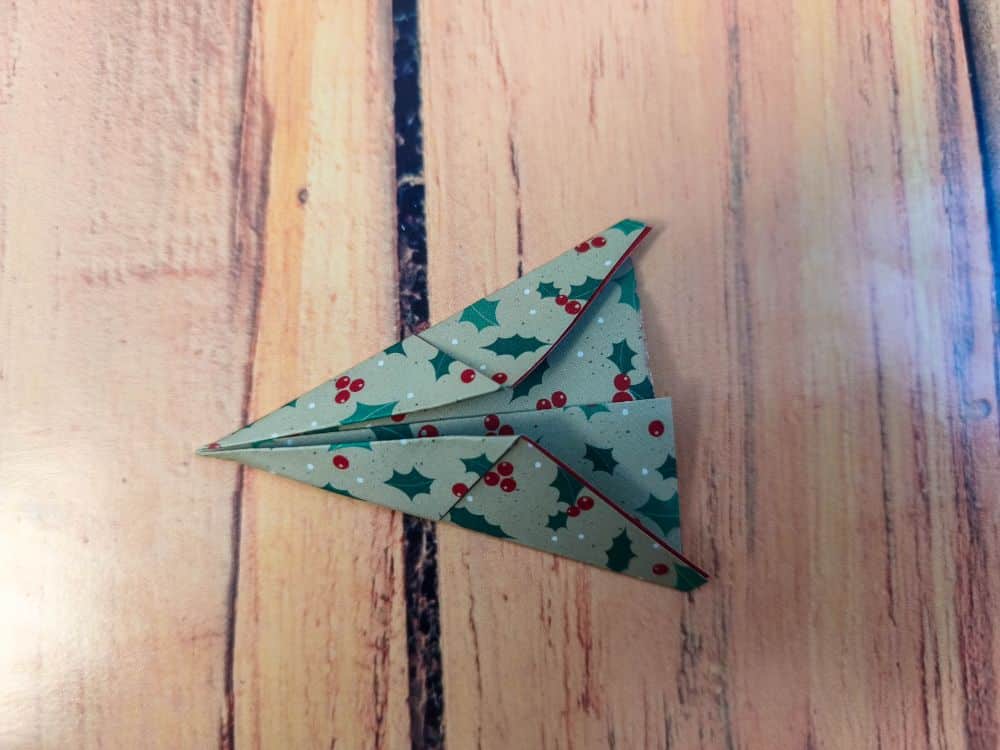 Ein Origami-Papierflugzeug aus weihnachtlichem Papier mit grünen Stechpalmenblättern und roten Beeren, das auf einer Holzunterlage ruht.