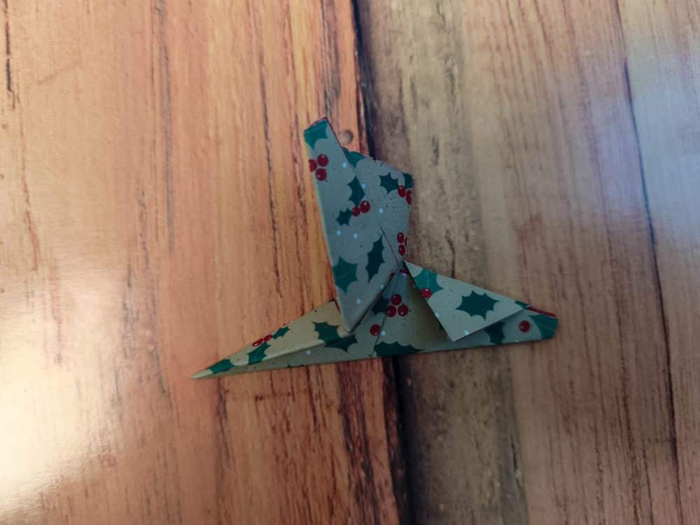 Ein kleines Stück Origamipapier, das in Form eines Vogels gefaltet ist, liegt auf einer Holzfläche. Das Origamipapier ist mit grünen Stechpalmenblättern und roten Beeren verziert.