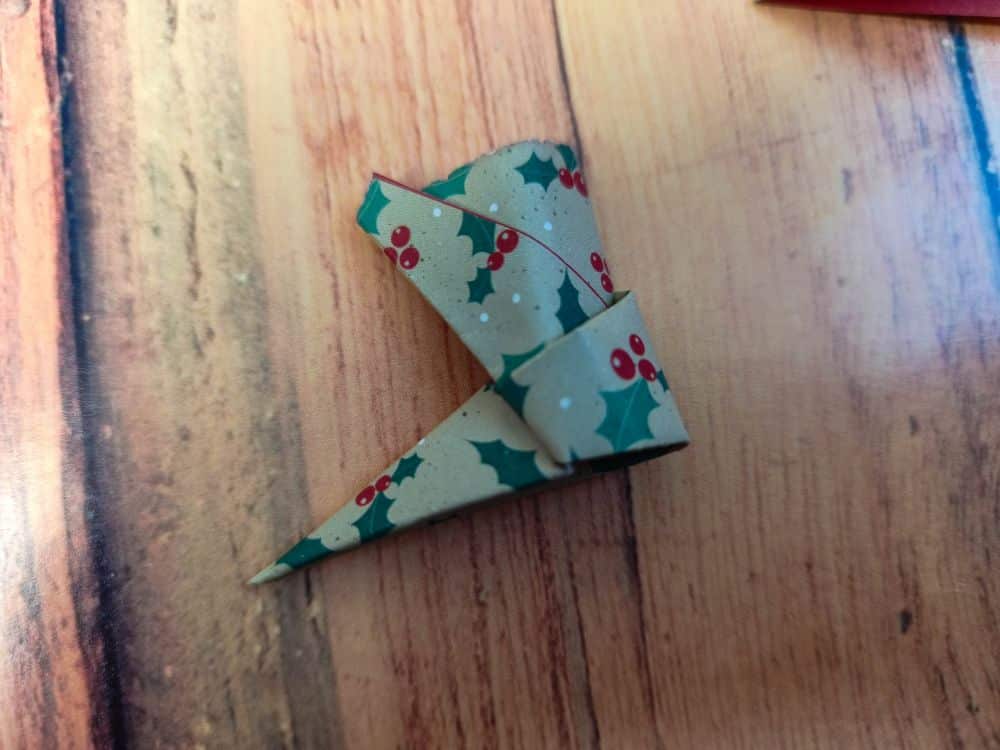 Ein gefaltetes Stück braunes Origamipapier mit einem weihnachtlichen Stechpalmen- und Beerenmuster, das auf einer Holzunterlage liegt. Das Papier ist zu einer spitzen, eckigen Form geformt.