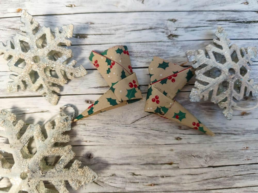 Zwei glitzernde weiße Schneeflocken und ein Paar Origami-Papierschlittschuhe mit Stechpalmen- und Beerenmotiven sind auf einer rustikalen Holzfläche angeordnet.