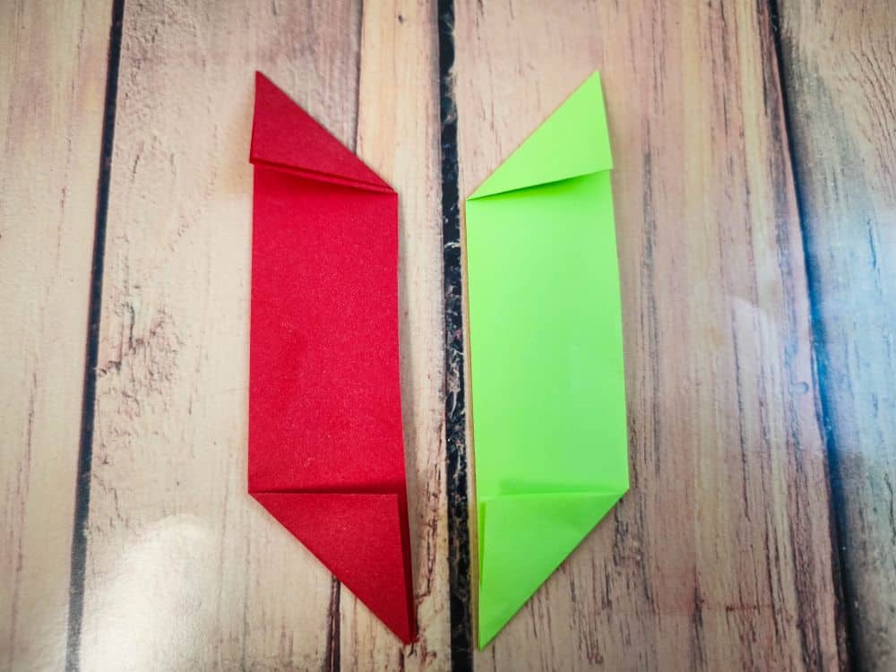 Zwei Origami-Papierformen, eine rote und eine grüne, werden nebeneinander auf eine Holzfläche gelegt. Jede Origami-Form hat spitze, nach innen gefaltete Ecken und ähnelt einem langen Parallelogramm.