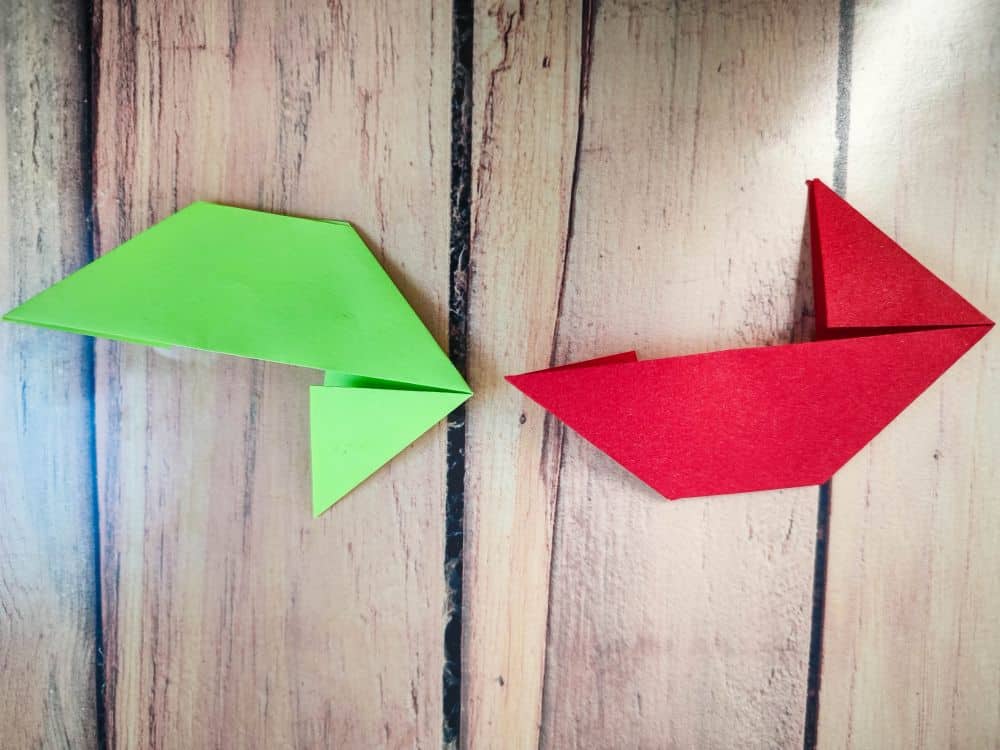 Zwei Origami-Papiervögel, ein grüner und ein roter, liegen nebeneinander auf einer Holzfläche. Die Origami-Vögel sind einfach und eckig gefaltet und zeigen in unterschiedliche Richtungen.