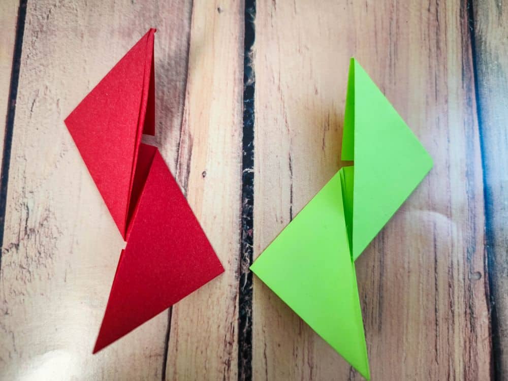 Zwei Origami-Formen, eine rote und eine grüne, die jeweils aus zwei verbundenen Dreiecken bestehen, werden nebeneinander auf eine Holzfläche gelegt.