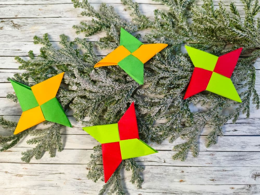 Vier bunte Origami-Ninja-Sterne in gelb-grüner und rot-grüner Farbkombination ruhen auf gefrosteten Kiefernzweigen auf einer Holzfläche.
