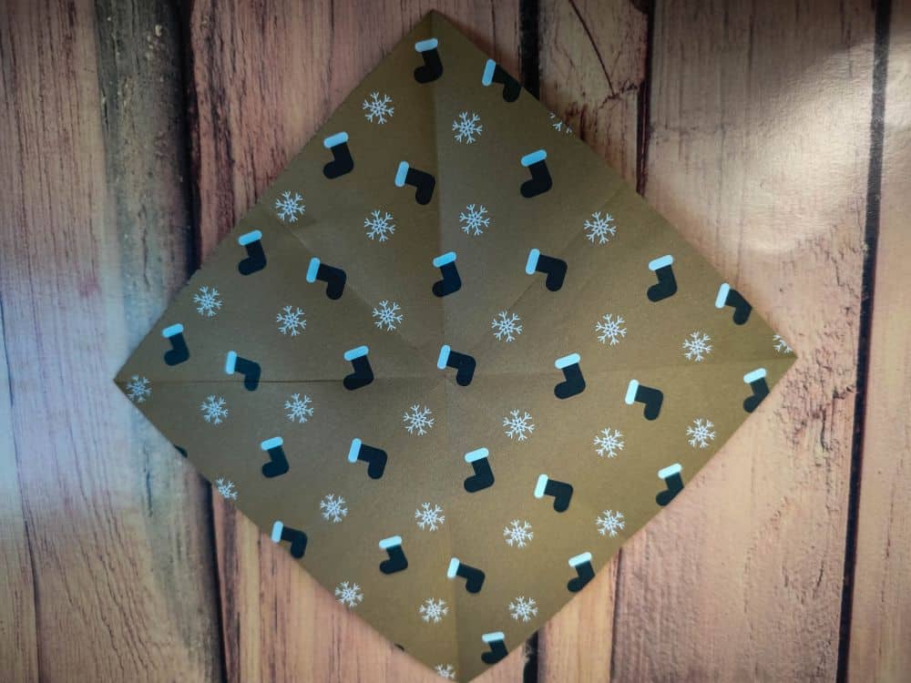 Ein quadratisches Stück braunes Origamipapier mit einem Muster aus schwarzen Weihnachtsstrümpfen und weißen Schneeflocken wird diagonal auf eine Holzfläche gelegt.