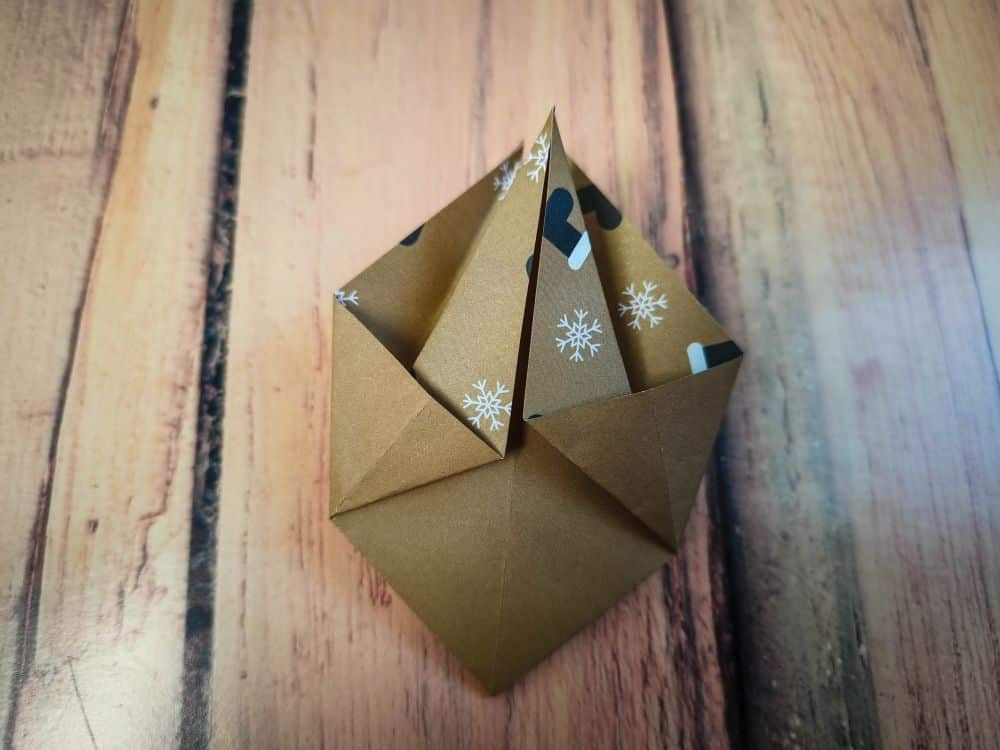 Eine Origami-Kreation aus braunem Papier mit weißen Schneeflockenmustern, die zu einer geometrischen, geschichteten Form gefaltet ist, liegt auf einer Holzfläche.