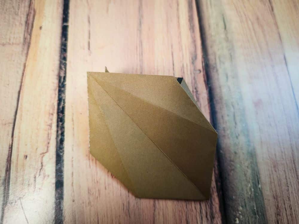 Eine Origami-Kreation mit scharfen Falten sitzt auf einer Holzoberfläche mit sichtbaren Maserungslinien und einer Mischung aus hellen und dunklen Holztönen.