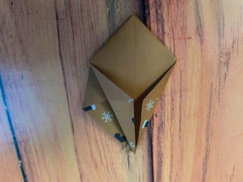 Ein braunes Origamipapier, das zu einer geometrischen, drachenähnlichen Form mit Schneeflockenmustern und schwarzen Herzmotiven gefaltet und auf eine holzstrukturierte Oberfläche gelegt wurde.