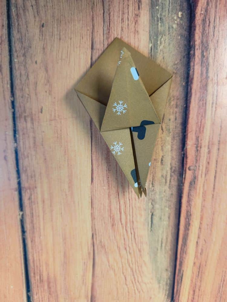 Eine braune Origami-Kreation mit weißen Schneeflockenmustern und einem kleinen schwarzen Fleck ruht auf einer Holzoberfläche. Das Origami ist gekonnt zu einer geometrischen Form mit spitzen Enden gefaltet.