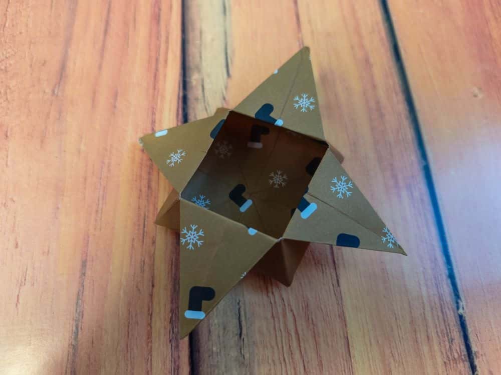 Eine braune, sternförmige Origamischachtel mit weißen Schneeflocken und schwarzen Formen liegt auf einer Holzfläche. Die Origamischachtel ist oben offen und offenbart ihre komplizierte innere Faltstruktur.
