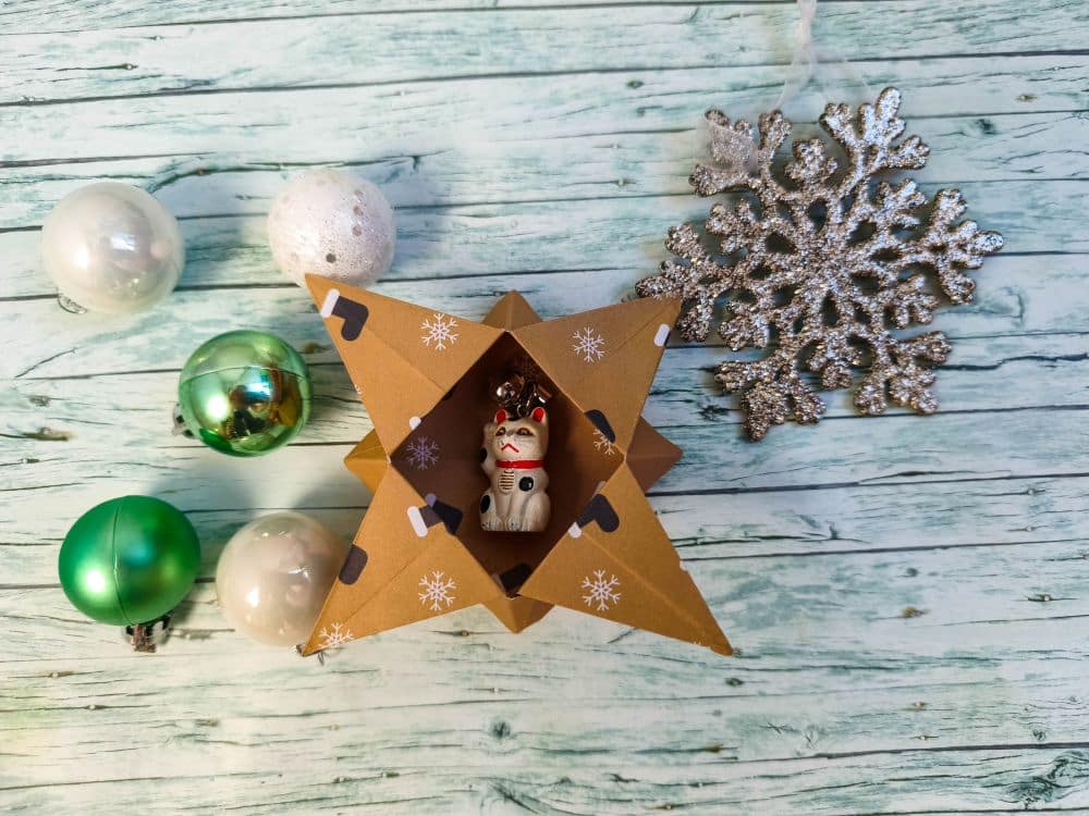 Eine kleine Glückskatze sitzt in einem sternförmigen Ornament aus Pappe mit Schneeflocken im Origami-Stil, umgeben von grünen und weißen Kugeln und einer glitzernden silbernen Schneeflocke auf einer hellen Holzoberfläche.