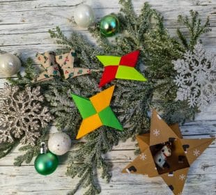 Ein festliches Arrangement mit grünen Tannenzweigen, silbernem Schneeflockenschmuck, grünen und weißen Kugeln, leuchtenden Origami-Sternen, einer gemusterten Papierschleife und einer Sternbox aus Pappe mit kleinen Tierfiguren.