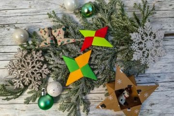 Ein festliches Arrangement mit grünen Tannenzweigen, silbernem Schneeflockenschmuck, grünen und weißen Kugeln, leuchtenden Origami-Sternen, einer gemusterten Papierschleife und einer Sternbox aus Pappe mit kleinen Tierfiguren.