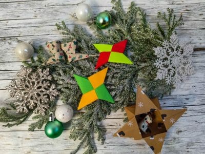 Ein festliches Arrangement mit grünen Tannenzweigen, silbernem Schneeflockenschmuck, grünen und weißen Kugeln, leuchtenden Origami-Sternen, einer gemusterten Papierschleife und einer Sternbox aus Pappe mit kleinen Tierfiguren.