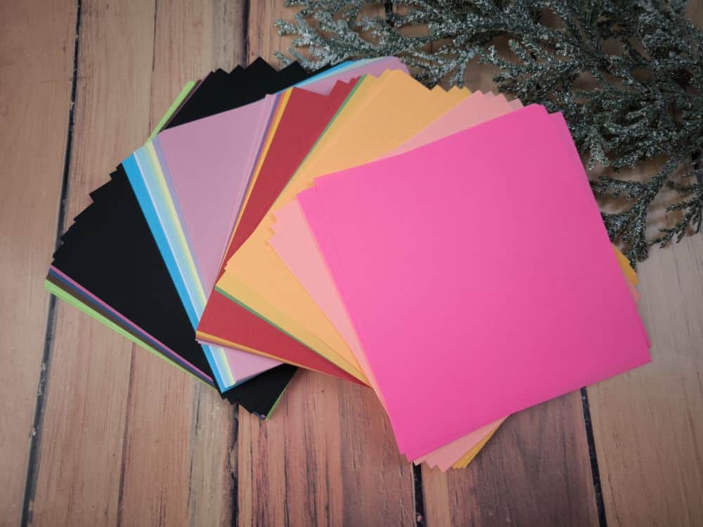 Ein Stapel bunter, quadratischer Origamipapiere, die fächerförmig auf einer hölzernen Oberfläche angeordnet sind, mit einer gefrosteten Pflanzendekoration in der Ecke - perfekt für Origami-Fans.