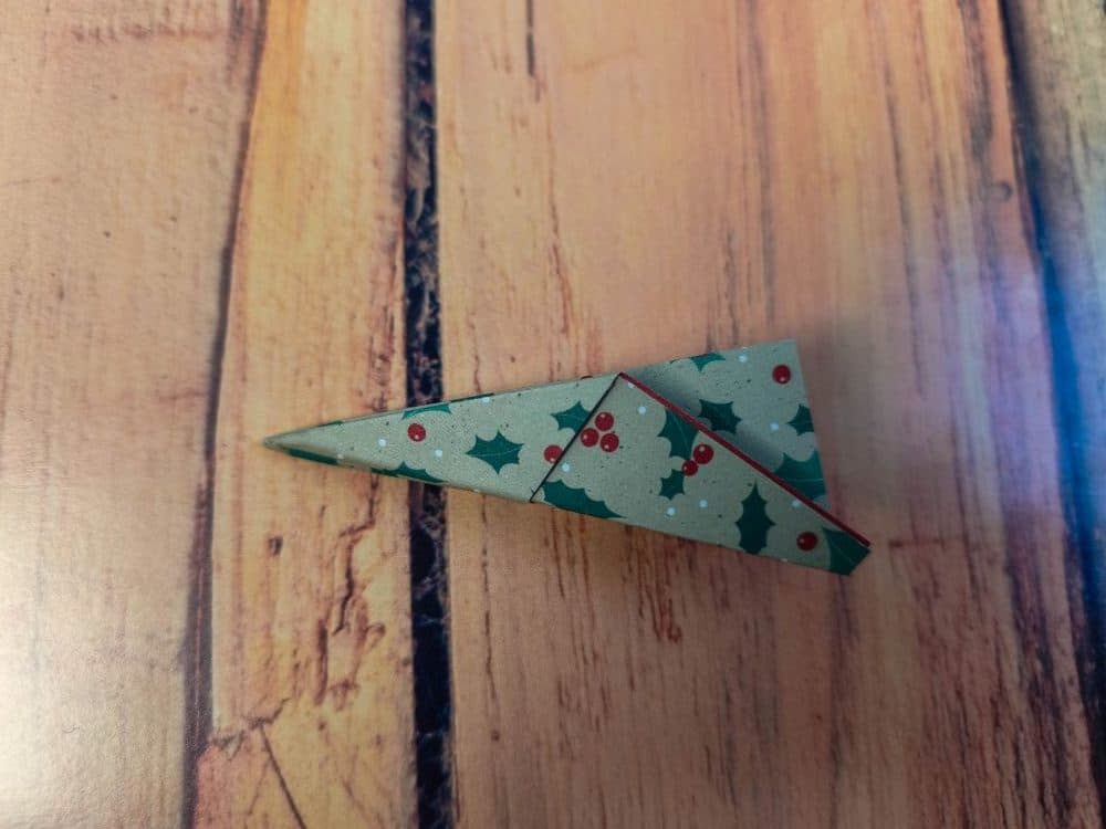 Ein kleines, dreieckiges Stück Papier mit einem Muster aus grünen Stechpalmen und roten Beeren liegt auf einer Holzfläche. Das Papier ist ordentlich im Origami-Stil gefaltet und bildet eine spitze Form.