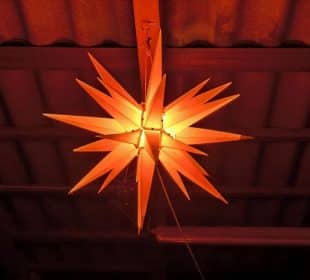 Von der Decke hängt eine leuchtende, sternförmige Laterne mit Stacheln, die ein warmes orangefarbenes Licht abgibt. Die gewellten Dachplatten und die sichtbaren Balken darüber verleihen dem Raum einen rustikalen Charme, der durch eine nahe gelegene Kadomatsu-Anlage ergänzt wird.