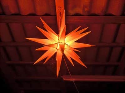 Von der Decke hängt eine leuchtende, sternförmige Laterne mit Stacheln, die ein warmes orangefarbenes Licht abgibt. Die gewellten Dachplatten und die sichtbaren Balken darüber verleihen dem Raum einen rustikalen Charme, der durch eine nahe gelegene Kadomatsu-Anlage ergänzt wird.