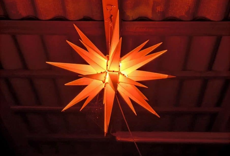 Von der Decke hängt eine leuchtende, sternförmige Laterne mit Stacheln, die ein warmes orangefarbenes Licht abgibt. Die gewellten Dachplatten und die sichtbaren Balken darüber verleihen dem Raum einen rustikalen Charme, der durch eine nahe gelegene Kadomatsu-Anlage ergänzt wird.